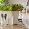 Jarra llenándose con agua del grifo junto a medidores de pH y EC y un cultivo hidropónico doméstico
