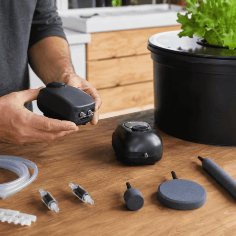 Persona comparando bombas de aire, tubos, válvulas y diferentes piedras difusoras junto a un sistema DWC doméstico